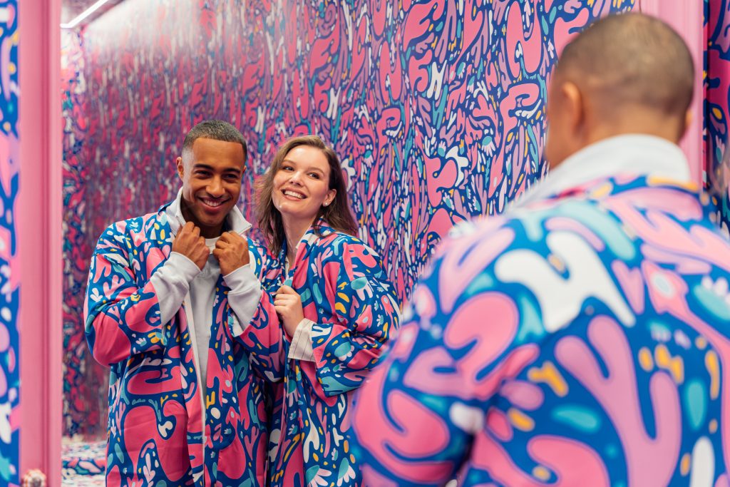 Two guests smiling in matching colorful robes in a vibrant mirrored IKONO room with pink and blue patterned walls.