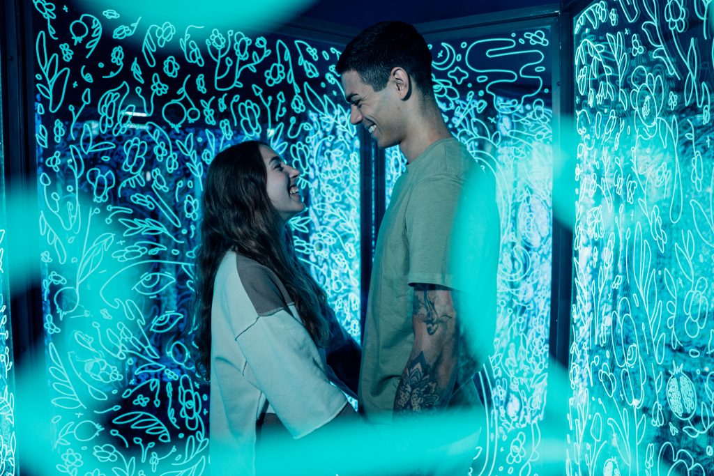 Two visitors smiling at each other inside a glowing turquoise room with illuminated doodle-style patterns on the walls.