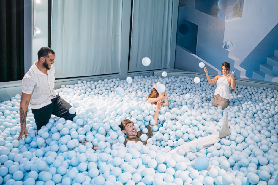  Four visitors playing and laughing in a large light blue ball pit inside an immersive IKONO room. 
