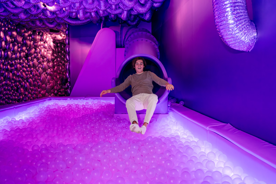 Visitor laughing as he slides into a glowing pink and purple ball pit inside an immersive IKONO play room.